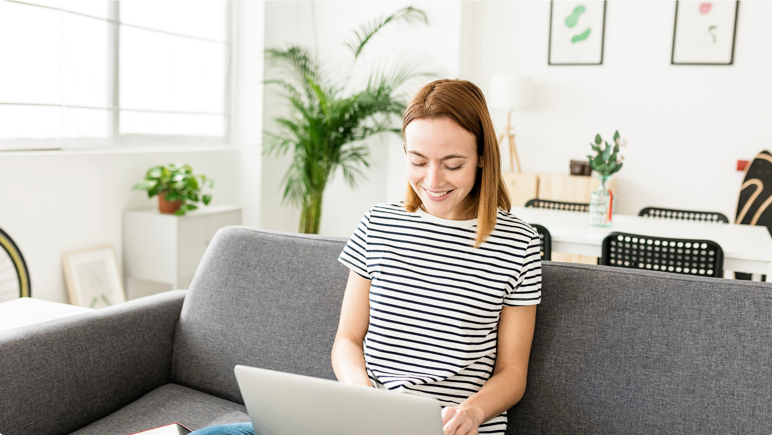 A young woman in a black and white striped top sits on a gray couch with an open laptop in her lap. She smiles as she types.