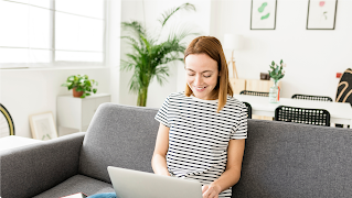 A young woman in a black and white striped top sits on a gray couch with an open laptop in her lap. She smiles as she types.