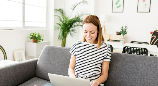 A young woman in a black and white striped top sits on a gray couch with an open laptop in her lap. She smiles as she types.