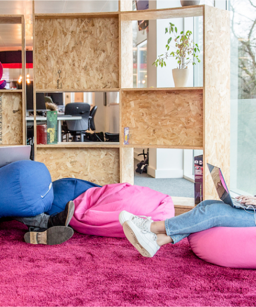 A man, on the left,  and a woman, on the right, sit on blue and pink bean bag chairs in the Octopus Energy office while working on laptops.