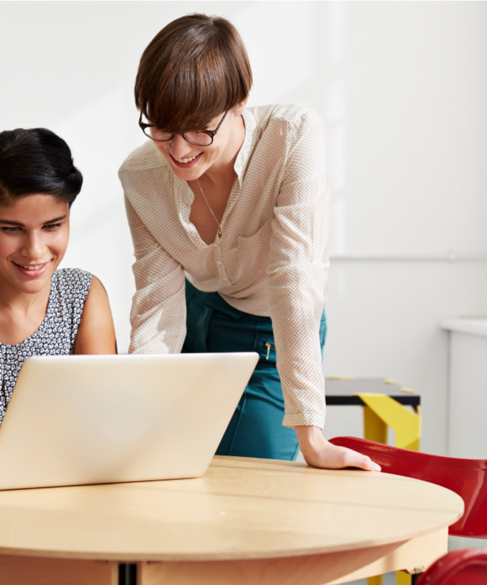 Two women smile and work together at a small round table with a laptop. One woman wears a patterned top and the other wears a white top.