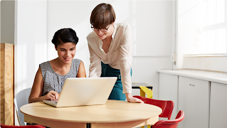 Zwei lächelnde Frauen arbeiten an einem kleinen runden Tisch mit einem Laptop. Eine trägt ein gemustertes Oberteil, die andere ein weißes.