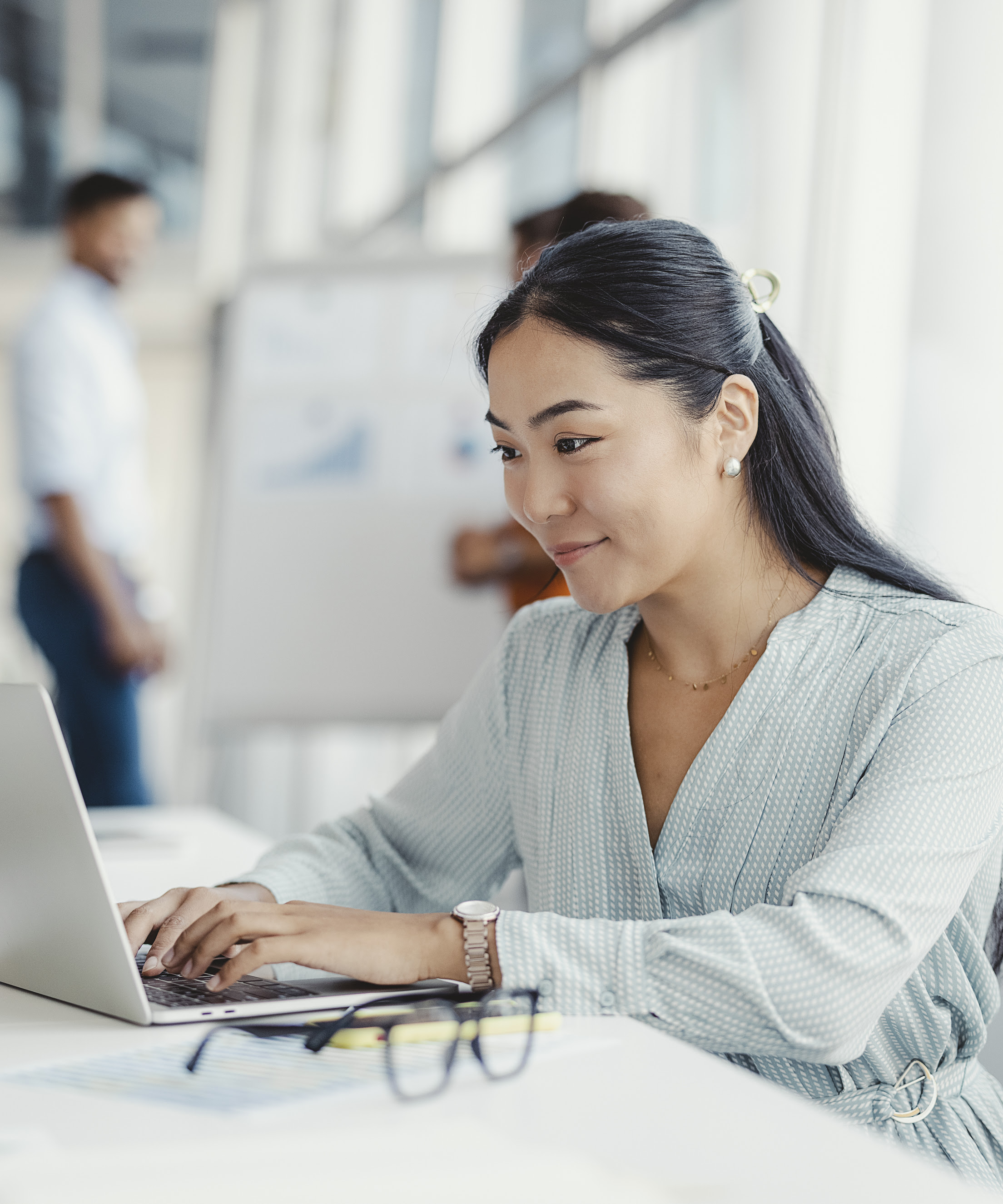 Businesswoman busy working on laptop computer at office with colleagues in the background.