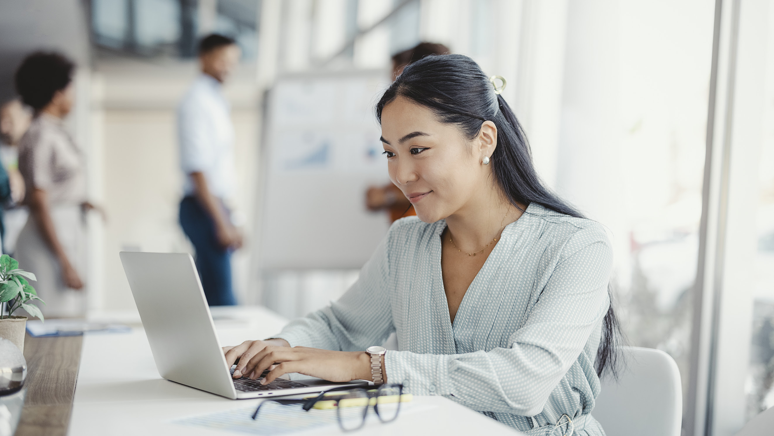 Businesswoman busy working on laptop computer at office with colleagues in the background.