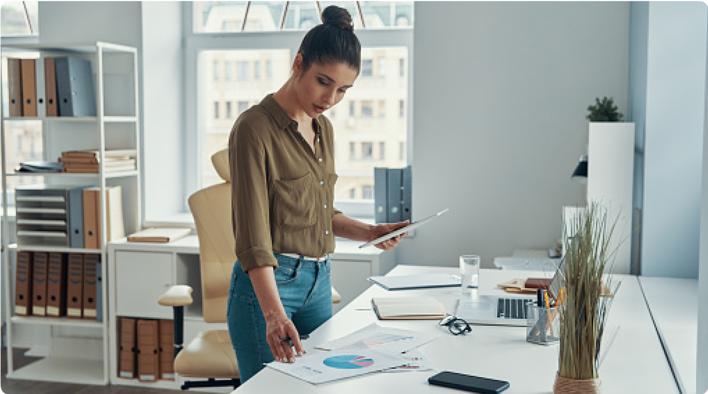 A young woman stands at a desk, with a tablet in her left hand, looking down at a printed pie chart to her right.