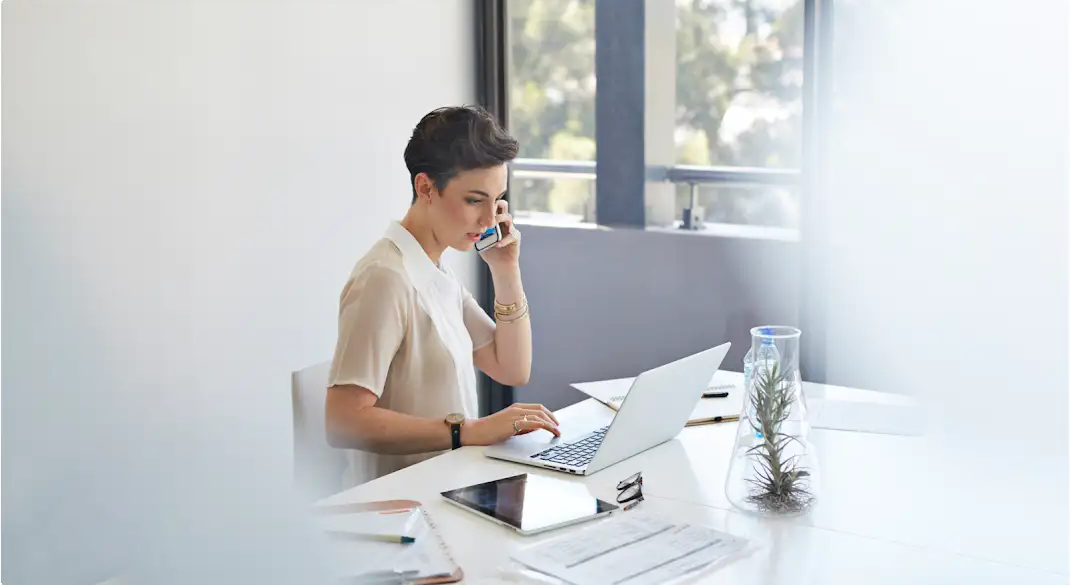 Eine Frau mit kurzen Haaren sitzt am Schreibtisch. Sie spricht in ihr Smartphone und hat einen Laptop sowie ein Tablet vor sich.