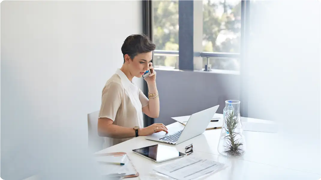 Eine Frau mit kurzen Haaren sitzt am Schreibtisch. Sie spricht in ihr Smartphone und hat einen Laptop sowie ein Tablet vor sich.