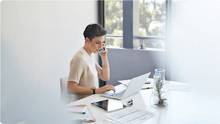 Eine Frau mit kurzen Haaren sitzt am Schreibtisch. Sie spricht in ihr Smartphone und hat einen Laptop sowie ein Tablet vor sich.