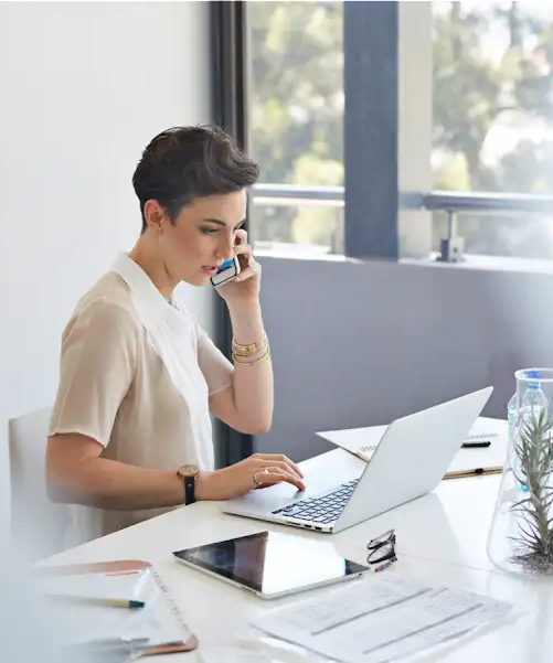 Eine Frau mit kurzen Haaren sitzt am Schreibtisch. Sie spricht in ihr Smartphone und hat einen Laptop sowie ein Tablet vor sich.