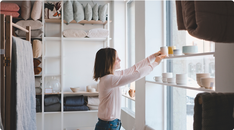 A business owner arranges products on a shelf in the front display window of her homegoods store.