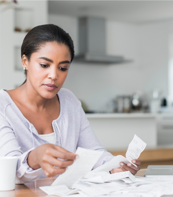 A woman sits at a table and sorts through a pile of receipts. She has a laptop to the left of the pile and a mug on the right.