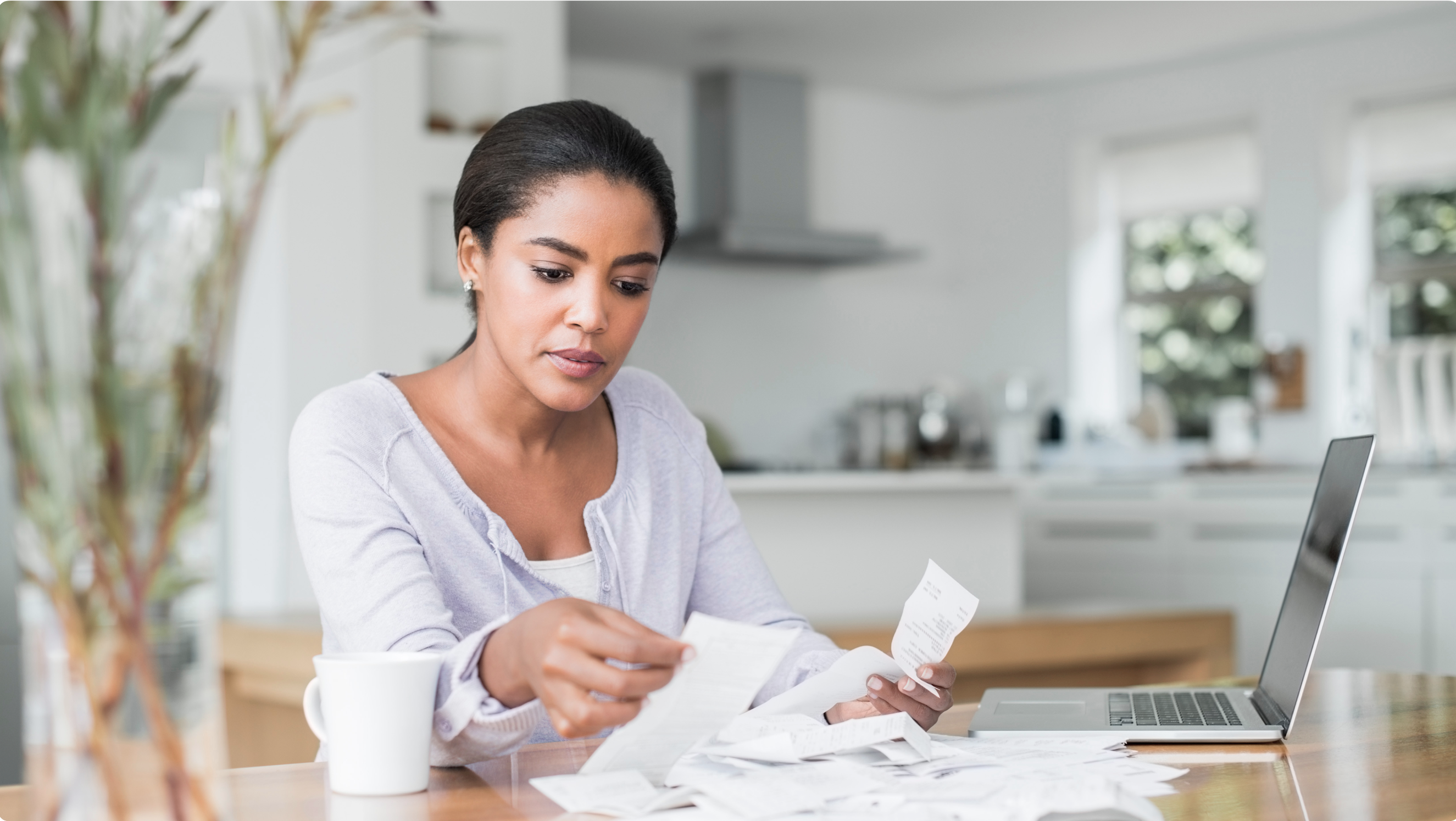 A woman sits at a table and sorts through a pile of receipts. She has a laptop to the left of the pile and a mug on the right.