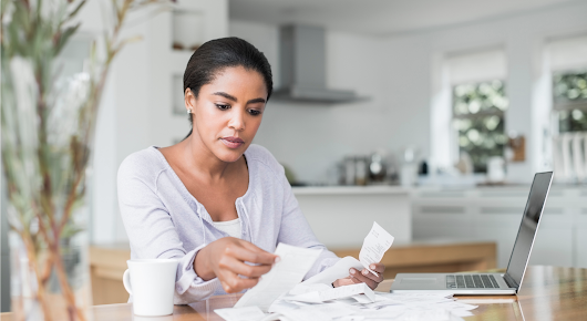 A woman sits at a table and sorts through a pile of receipts. She has a laptop to the left of the pile and a mug on the right.