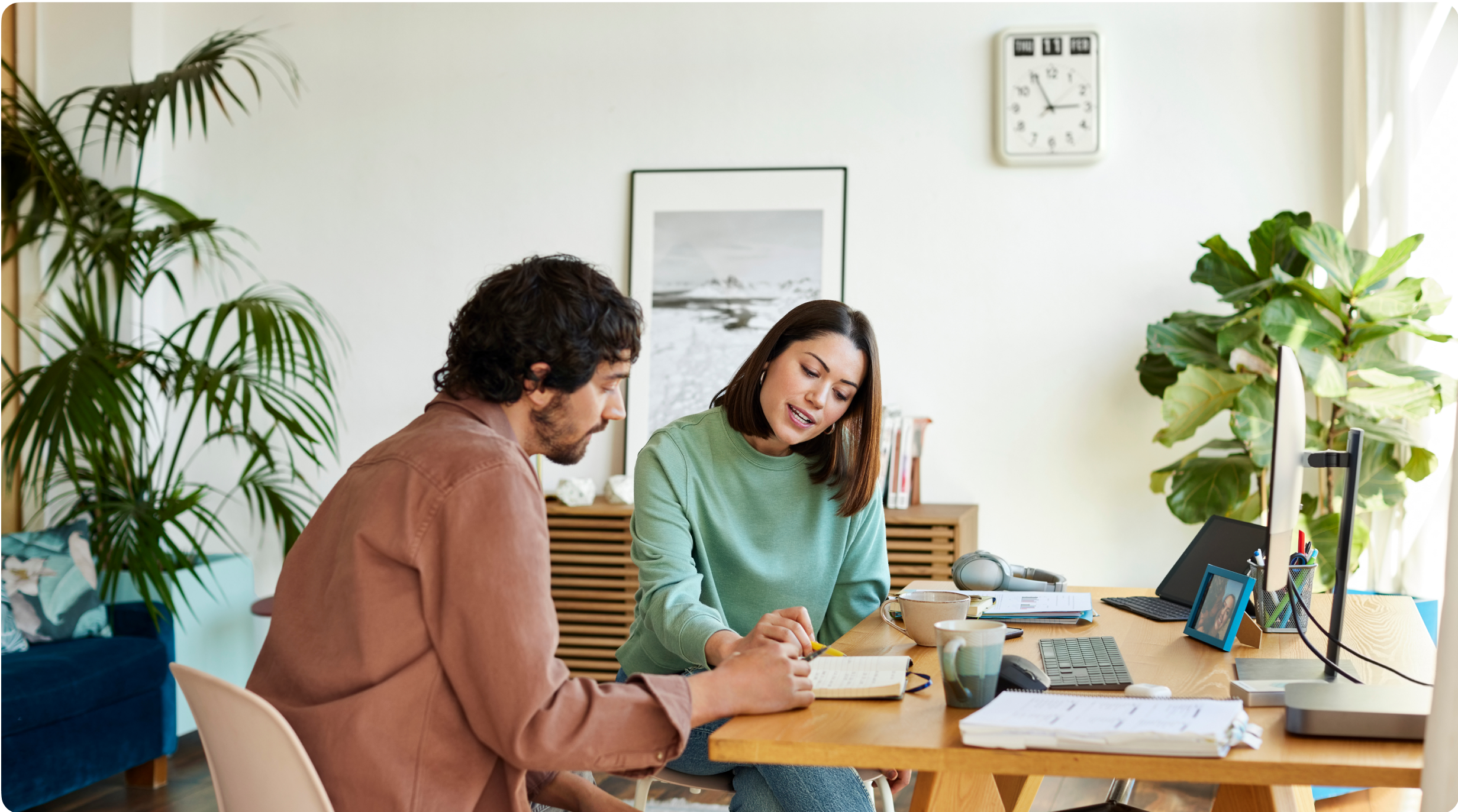 A man and woman sit at a desk, in a home office, reviewing notes together. The woman points to a page and the man follows along.