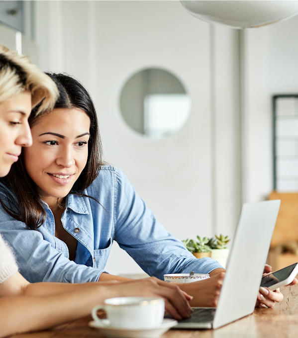 Two women use a laptop. One, in a white top, types while the other, in a blue top, holds a smartphone and looks at the laptop screen.