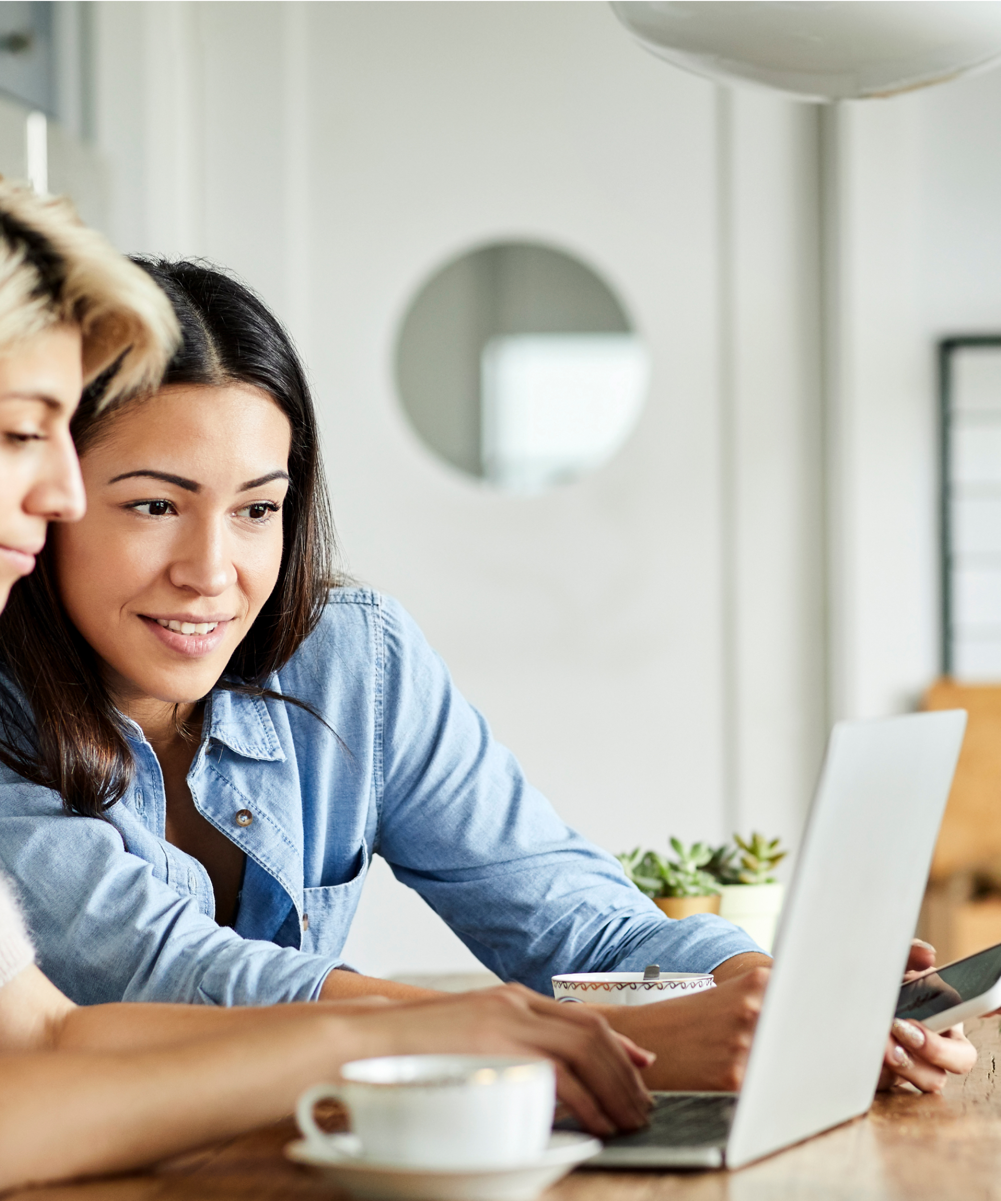 Two women use a laptop. One, in a white top, types while the other, in a blue top, holds a smartphone and looks at the laptop screen.