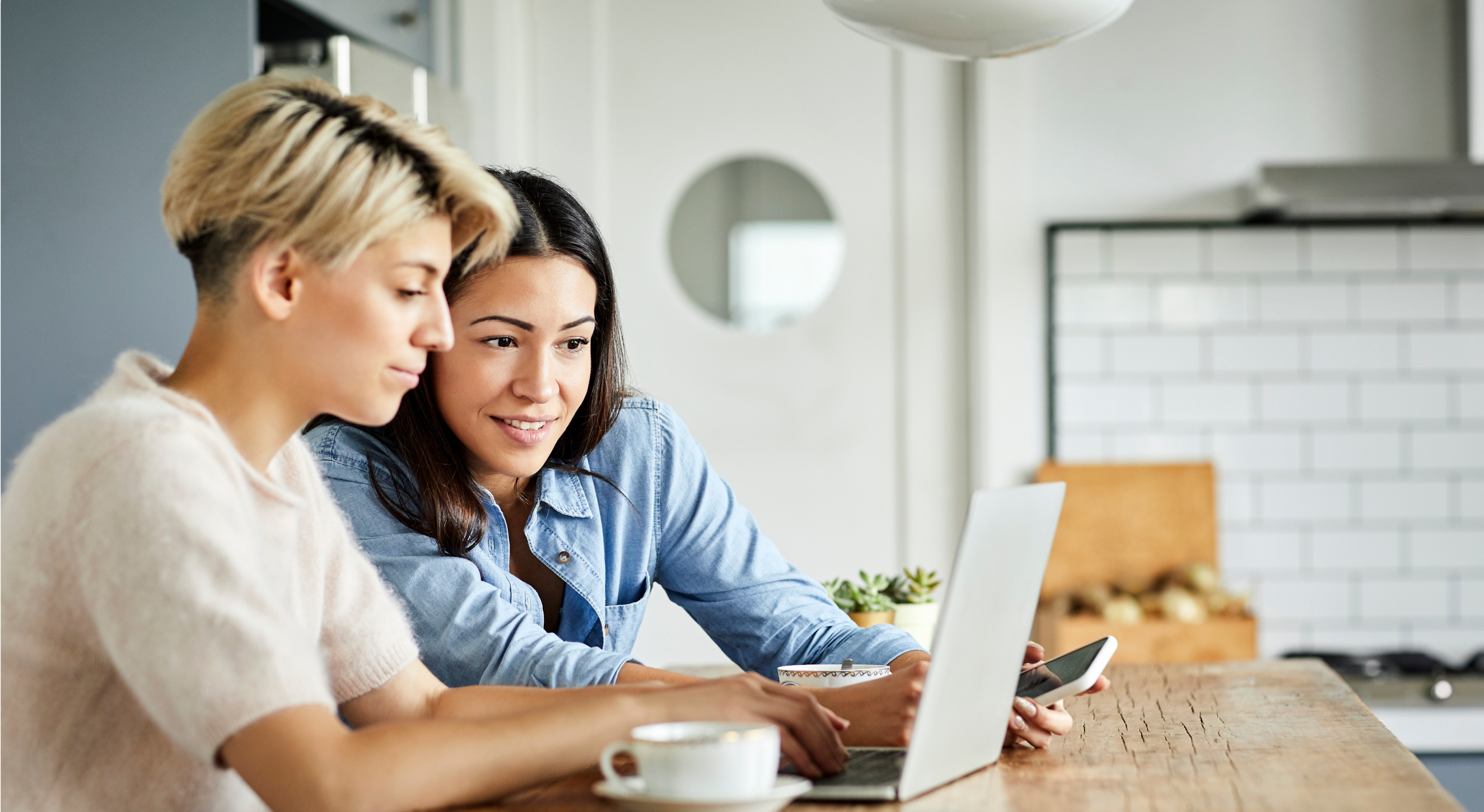 Two women use a laptop. One, in a white top, types while the other, in a blue top, holds a smartphone and looks at the laptop screen.