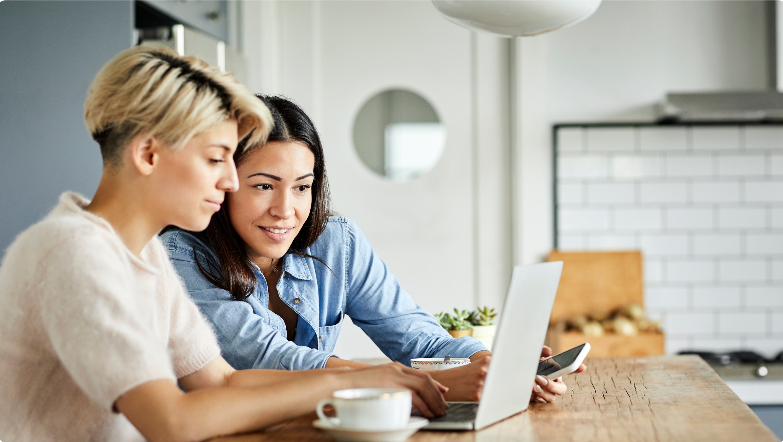 Two women use a laptop. One, in a white top, types while the other, in a blue top, holds a smartphone and looks at the laptop screen.