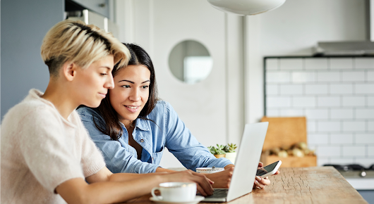 Two women use a laptop. One, in a white top, types while the other, in a blue top, holds a smartphone and looks at the laptop screen.