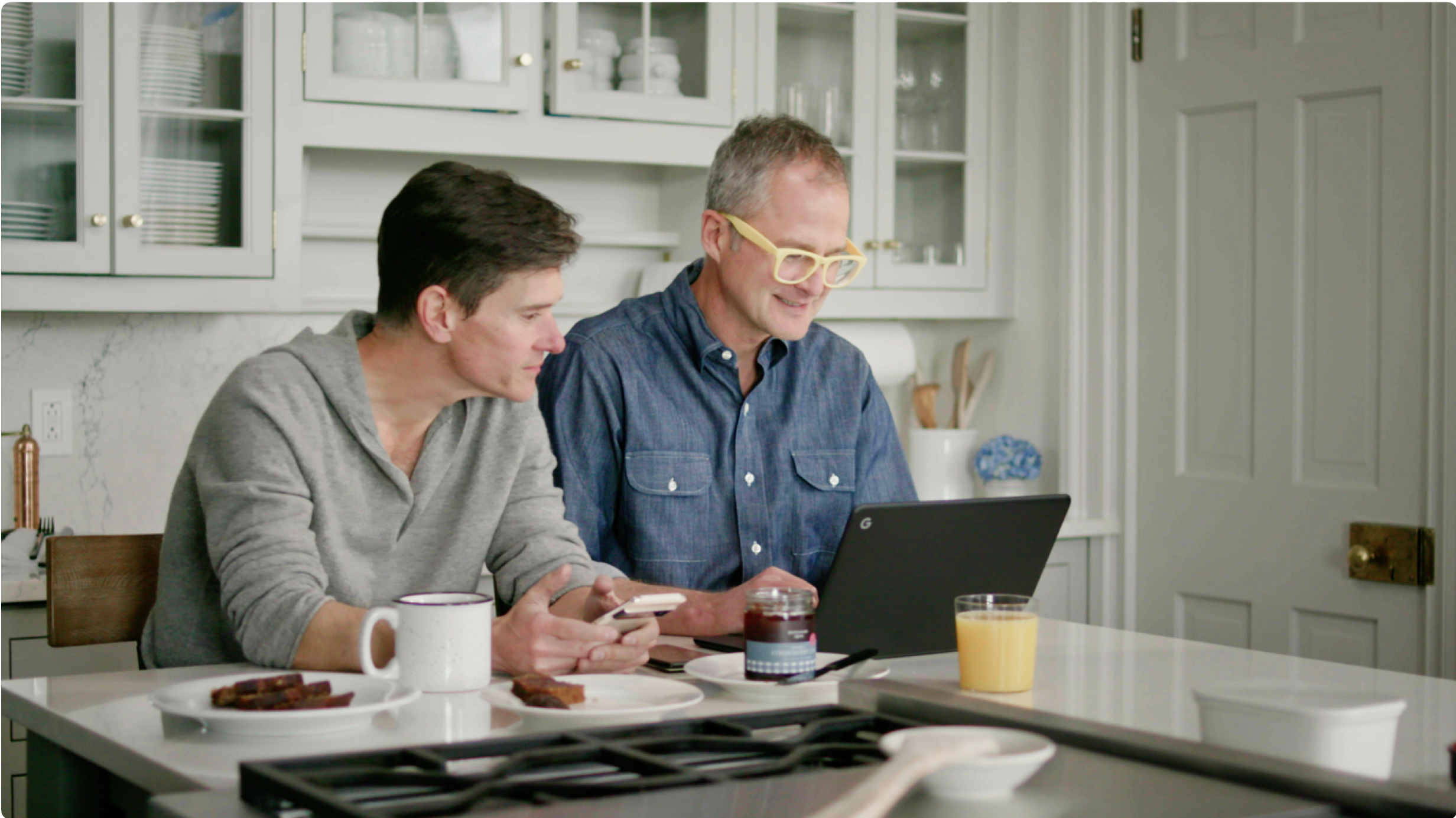 Brent and Josh work together at a kitchen island. Drinks and plates of food are scattered around them.