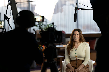 Behind the scenes shot of a woman in a white V-neck sweater smiling while sitting and speaking to camera.