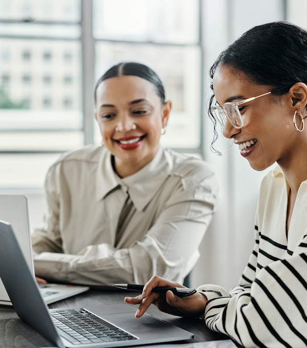 Two women smile as they collaborate at a desk while on their computers.