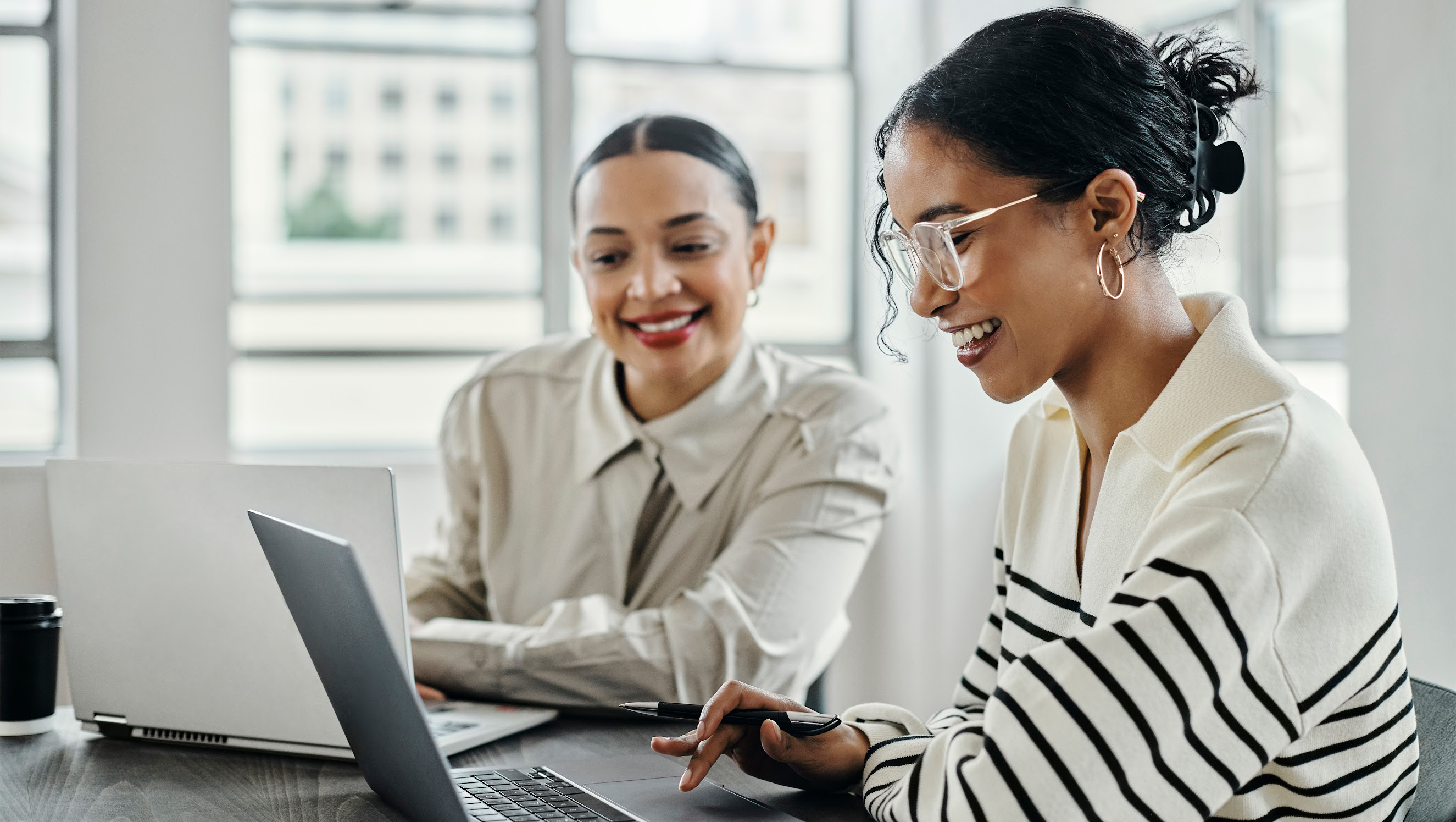 Two women smile as they collaborate at a desk while on their computers.