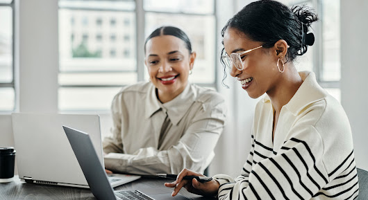 Two women smile as they collaborate at a desk while on their computers.