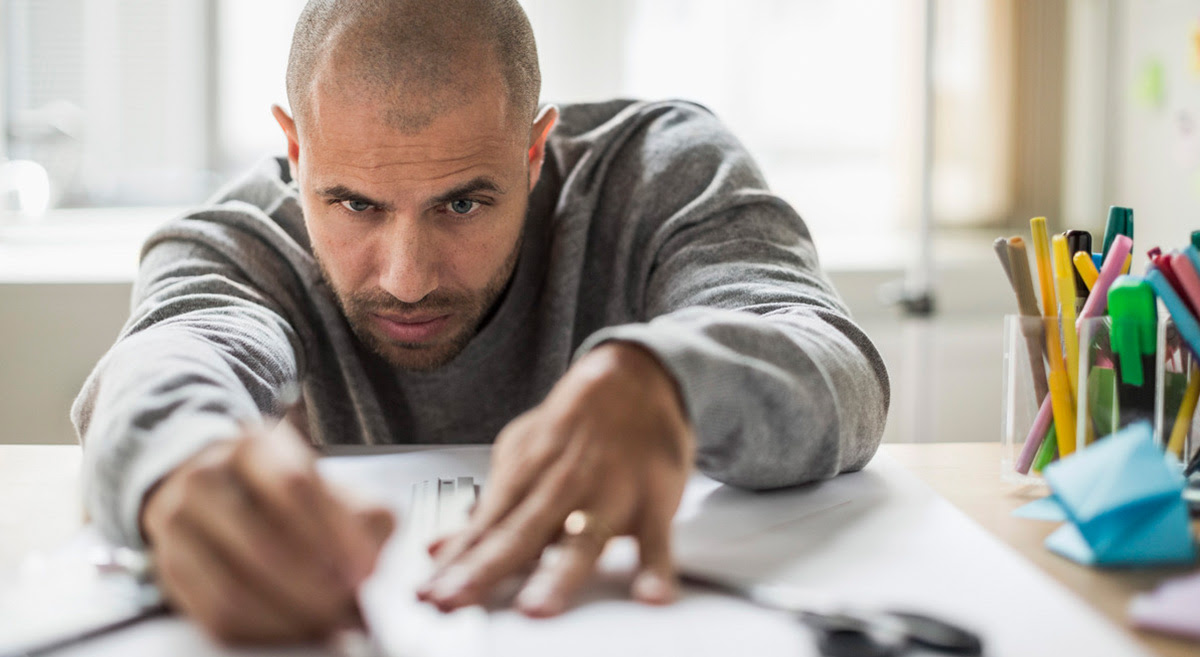 A man working with ruler and pen on a table with a large sheet of white paper and some cups of colorful pens