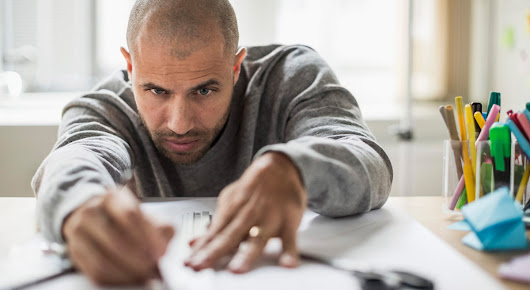 A man working with ruler and pen on a table with a large sheet of white paper and some cups of colorful pens