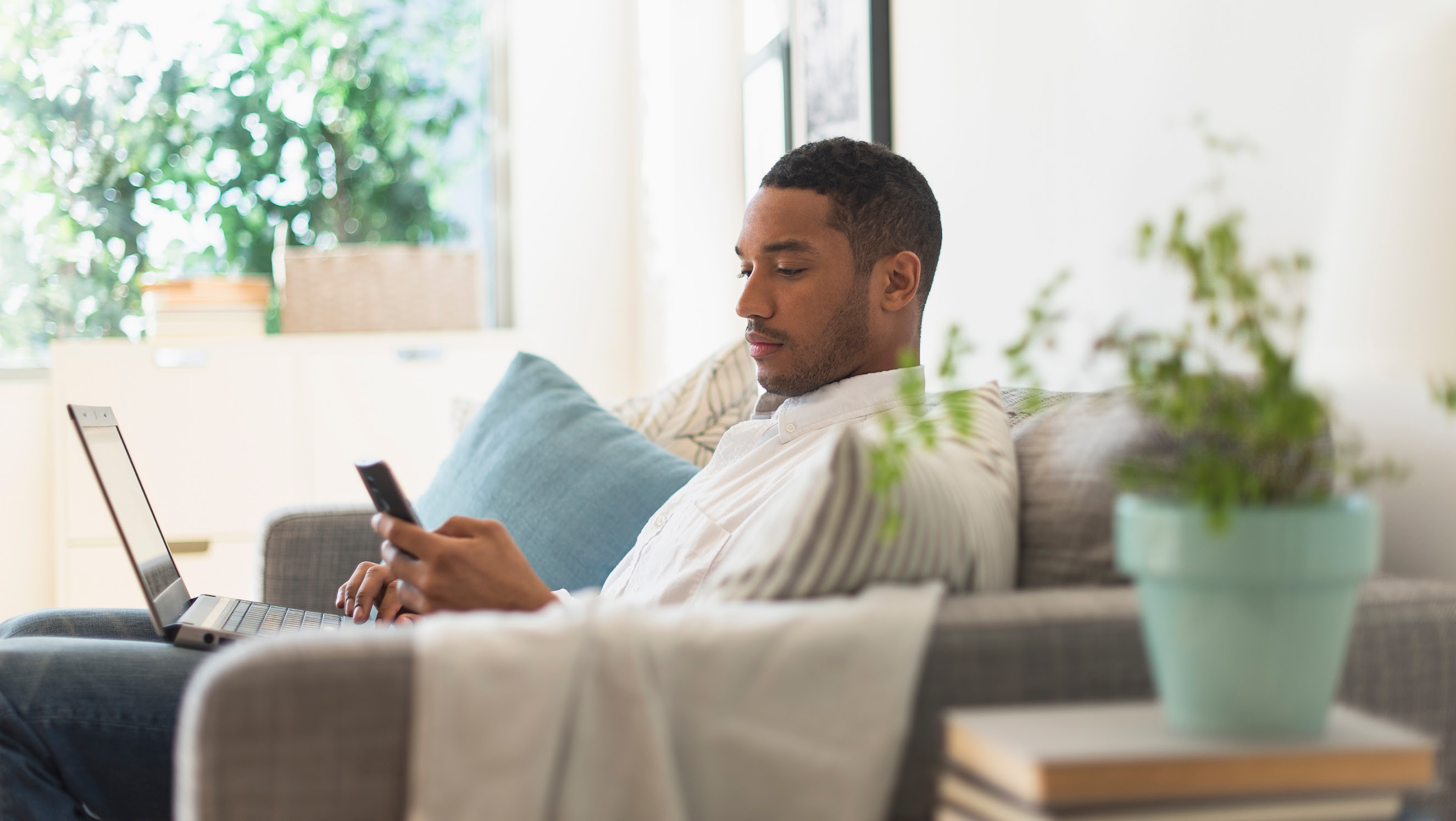 Man sitting on couch with a laptop in his lap and a mobile phone in his hand