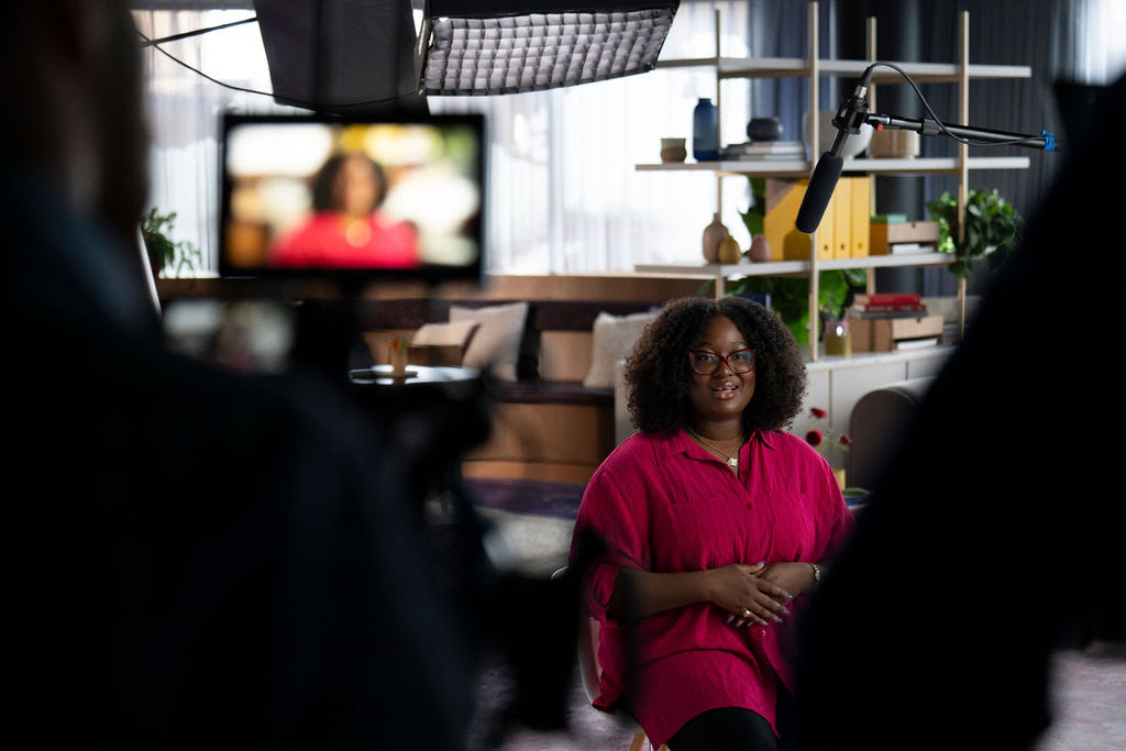 Behind the scenes shot of a woman with glasses in a red button-up shirt speaking to camera on a set.