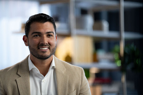 Man in a white button-up shirt and a tan blazer smiling and speaking to camera against a blurred background.