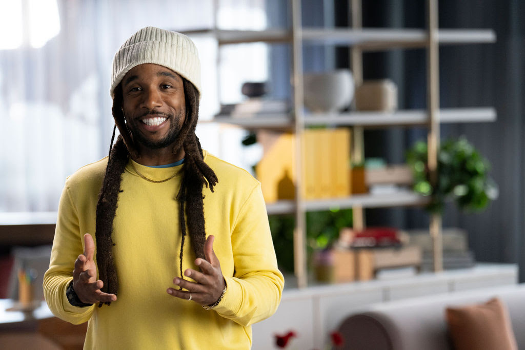 Man in a beanie and yellow sweater smiles while speaking animatedly to camera against a blurred background.