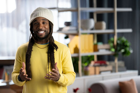 Man in a beanie and yellow sweater smiles while speaking animatedly to camera against a blurred background.