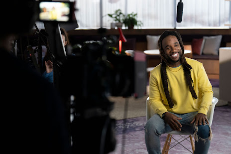 Behind the scenes shot of a man in a yellow shirt smiling and sitting while speaking to camera.