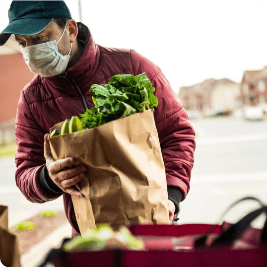 A 412 Food Rescue employee wearing a green cap and a disposable face mask holds a paper grocery bag containing lettuce, bananas, and other foodstuff; before him, a graphic reads, “50% cut down of reporting time”