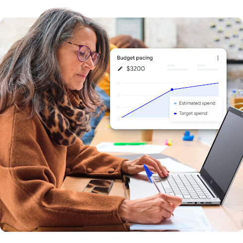 A woman, wearing glasses, works on a laptop and writes on a notepad. A line graph depicting a budget floats to her right.