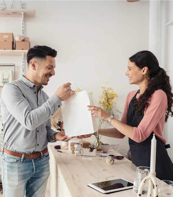 A woman behind a shop counter hands a shopping bag to a man who bought something from the store.