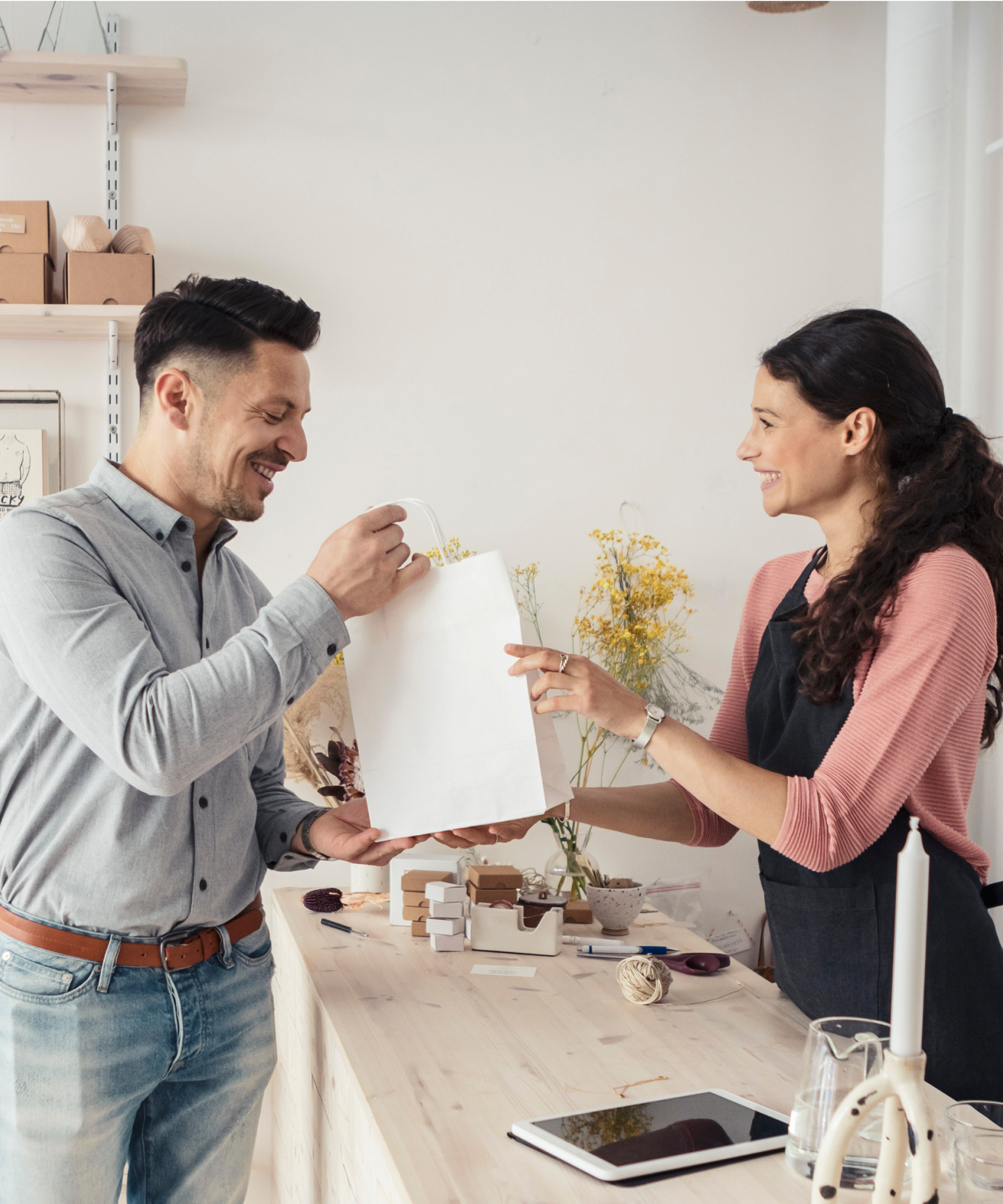 A woman behind a shop counter hands a shopping bag to a man who bought something from the store.