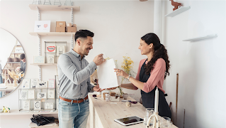 A woman behind a shop counter hands a shopping bag to a man who bought something from the store.