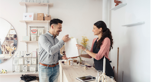 A woman behind a shop counter hands a shopping bag to a man who bought something from the store.