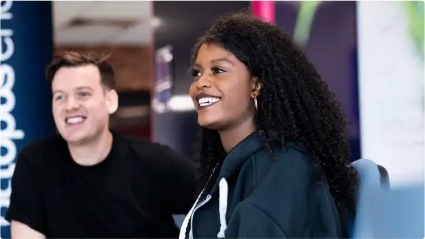 A man in a black shirt sits next to a woman wearing a blue hoodie in the Octopus Energy office. They smile while looking to the left of the camera.