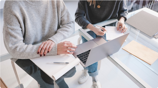 Two people work while sitting next to each other at a glass desk with a laptop.