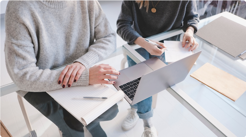 Two people work while sitting next to each other at a glass desk with a laptop.