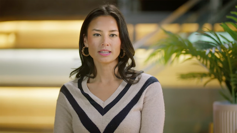 A woman with dark, shoulder-length hair speaks while wearing a beige v-neck sweater with a navy blue chevron pattern