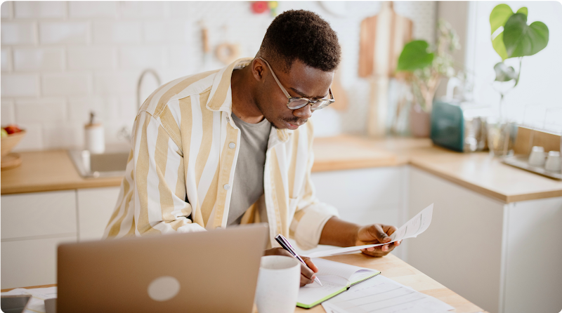 A young business owner stands in his kitchen as he  reviews paperwork and jots down notes in front of a laptop.