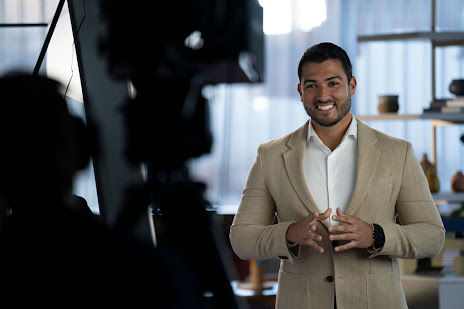 Behind the scenes shot of a man in a white button-up shirt and a tan blazer speaking animatedly to camera against blurred background.