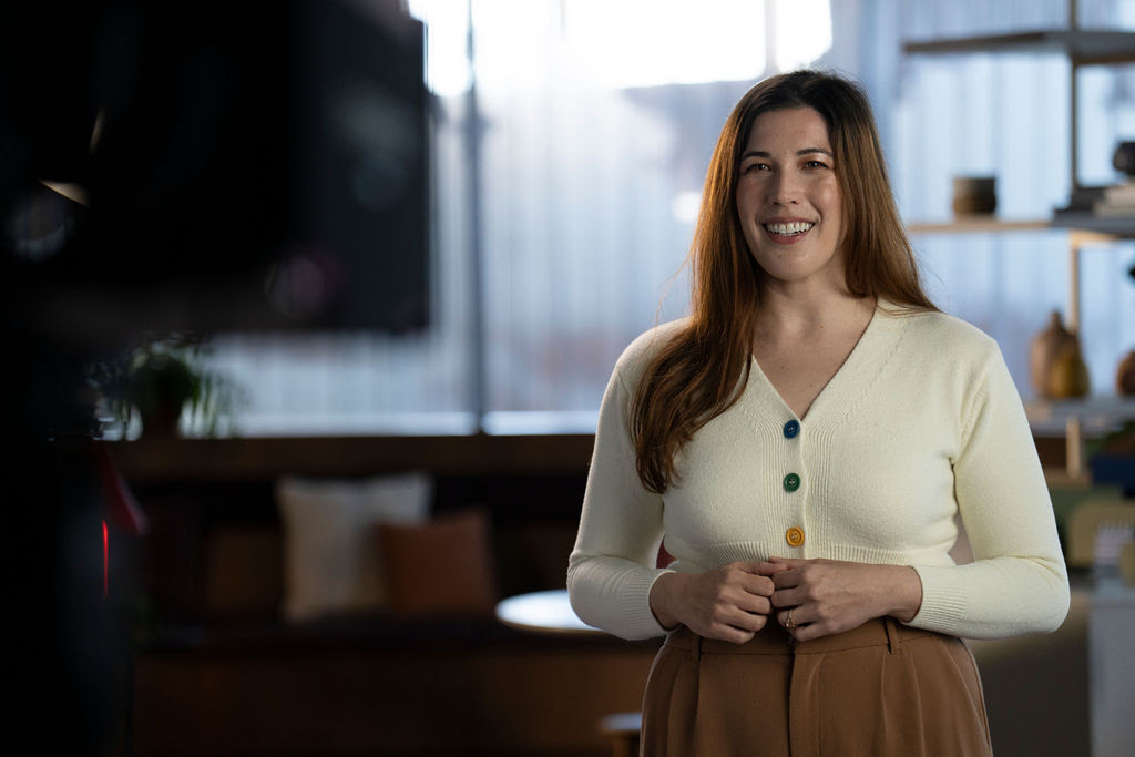 Behind the scenes shot of a woman in a white V-neck sweater smiling while speaking to camera against a blurred background.