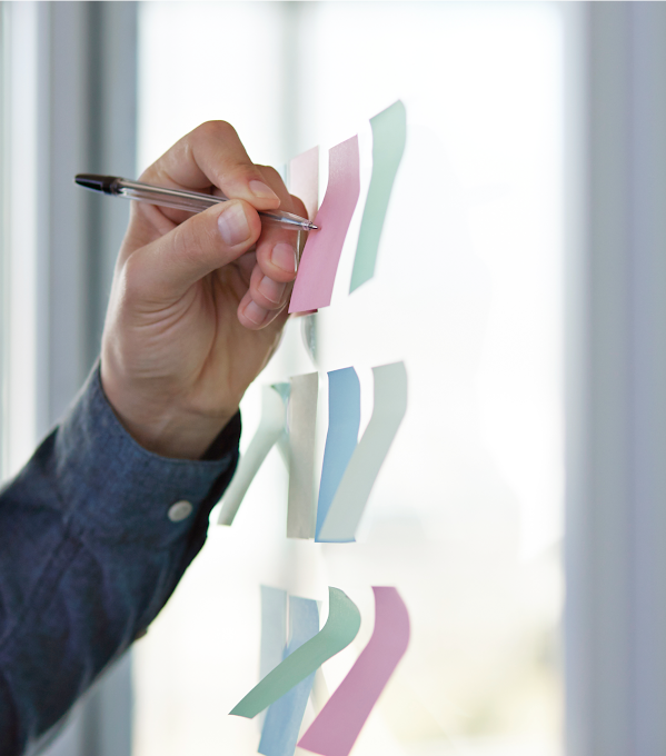 A person’s left arm writes on sticky notes that are placed on a glass wall.